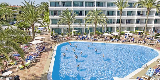 Ein ovalförmiger Pool befindet sich vor einem mehrstöckigen Gebäude umgeben von Palmen. Im Pool stehen Menschen und machen Gymnastikübungen.