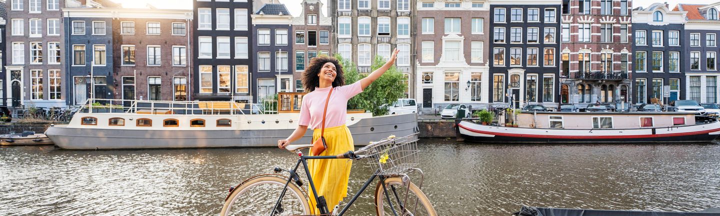 Eine junge Frau steht freudig mit ihrem Fahrrad an einer Gracht in Amsterdam. Sie reist die Arme hoch und lacht, im Hintergrund eine typisch niederländische Häuserreihe.