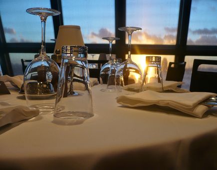 Ein gedeckter Tisch mit weißer Tischdecke. Im Hintergrund mit großen Fenster und Ausblick auf das Meer.