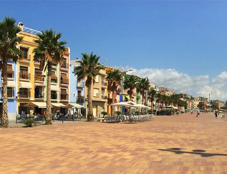 An einer bepflasterten Strandpromenade stehen Palmen, dahinter Restaurants mit Außenplätzen und Markisen und bunten Häuserfassaden.
