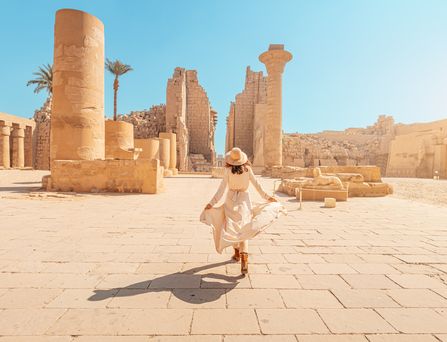 Eine in beige gekleidete Frau mit Sonnenhut läuft durch eine Ruinenlandschaft. Der Himmel ist strahlend blau.