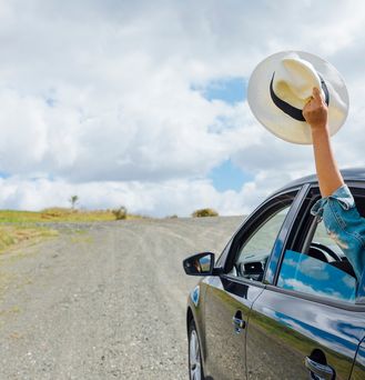Ein Auto fährt auf einer Straße und aus dem Rückfenster hebt eine Hand einen Sonnenhut in die Luft.