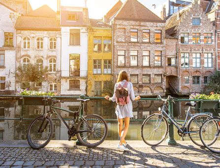 Eine junge Frau steht in Sommerkleidung mit Rucksack zwischen zwei Fahrrädern und blickt auf eine Gracht.