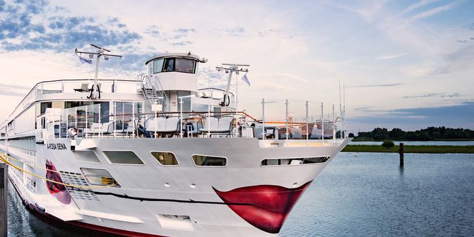 Weißes Passagierschiff mit rotem Rosenmuster am Bug auf ruhigem Gewässer unter bewölktem Himmel.