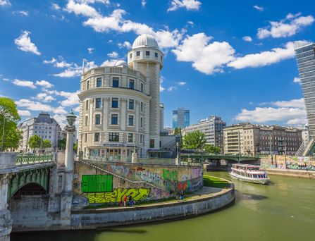 An einem grünen Fluss stehen historische und moderne Gebäude und ragen in den blauen Himmel. Am Rand einer schönen Brücke befindet sich eine mit Grafitti bemalte Wand.