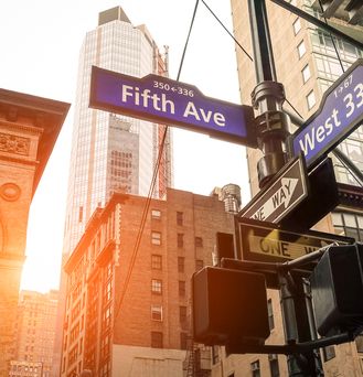 Eine Straßenkreuzung in New York. Auf einem Schild steht "Fifth Ave" und "West 33rd St". Die tiefstehende Sonne scheint durch die Wolkenkratzer.