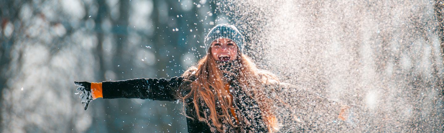 Eine junge blonde Frau mit Mütze und Handschuhen breitet lachend die Arme aus und wirbelt pulvrigen Schnee hoch.