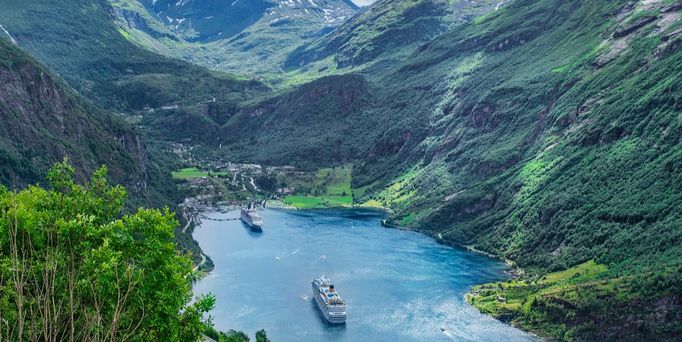 Inmitten eines norwegischen, saftgrünen Fjords befinden sich 2 Hochseekreuzfahrtschiffe. EIns liegt am Hafen an und ein weiteres bewegt sich von ihm weg.