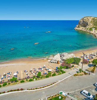 Direkt an einer felsigen Bucht am Meer liegt eine Wiese mit grünen Bäumen und ein Pool mit vielen Sonnenliegen mit Schirmen.