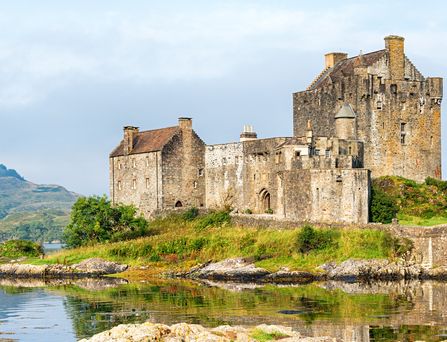 Eine mittelalterliche, etwas heruntergekommene Burg steht auf einem grünen, kleinen Hügel, welcher von einem See umringt ist.