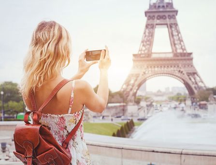 Eine junge, blonde Frau in Sommerkleidung steht vorm Eifelturm und hält ihr Handy zum fotografieren in den Händen.