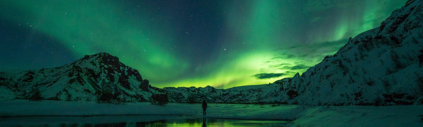 Der dunkel Nachthimmel wird von grünen Polarlichtern durchzogen. Eine Person steht, mit dem Rücken zugewandt und Wanderstöcken im weißen Schnee. Dahinter schneebedeckte Berge.