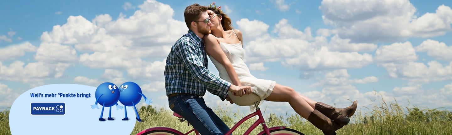 Kurzurlaub Valentinstag: Ein Mann fährt mit einem roten Fahrrad durch eine grüne Landschaft und eine Frau sitzt im weißen Sommerkleid auf dem Lenker