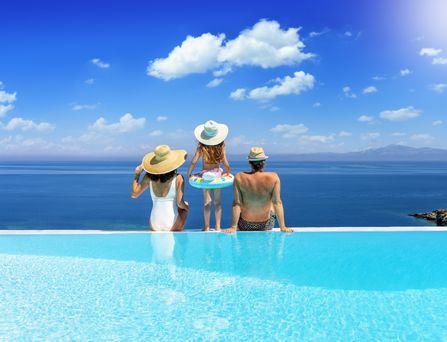 Eine Frau, ein Mann und ein kleines Mädchen in Badesachen und Sonnenhüten sitzen bzw. stehen am Rand eines hellblauen Infinity-Pools. Sie blicken hinab auf das tiefblaue Meer.