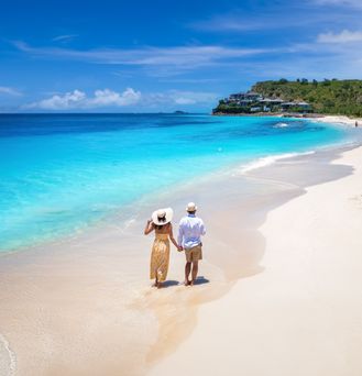 Ein Paar in Sommerkleidung läuft Hand in Hand an einem weißen Sandstrand entlang. Das Wasser ist türkisblau und im Hintergrund tropischer Wald und einzelne Häuser.