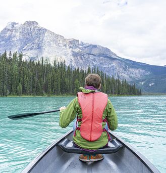 Ein Mann in Outdoor-Kleidung sitzt in einem Boot mit einem Paddel in der Hand. Im Hintergrund Berge und Wald.