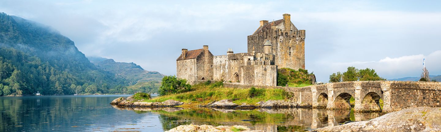 Eine mittelalterliche, etwas heruntergekommene Burg steht auf einem grünen, kleinen Hügel, welcher von einem See umringt ist.