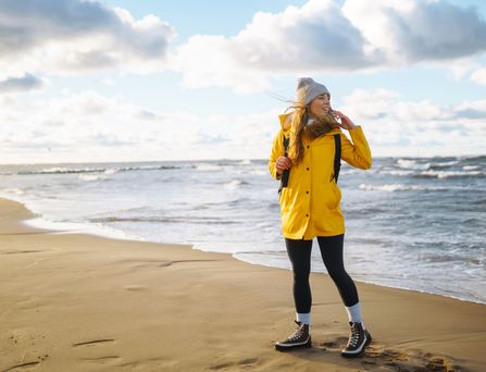 Eine junge Frau mit gelber Regenjacke und grauer Mütze steht am menschenleeren Strand. Der Wind weht ihr durch die Haare und sie blickt aufs Meer.