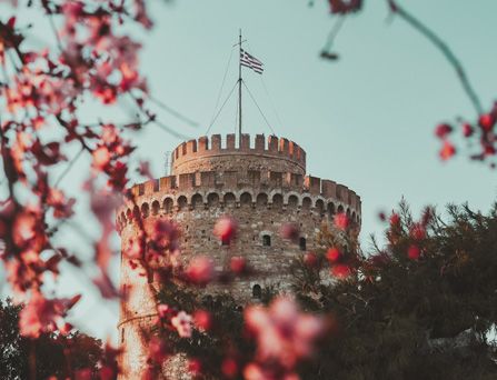 Eine mittelalterlicher Burgturm mit Griechenlandflagge ragt in den hellblauen Himmel. Im Vordergrund pinke Blumen.