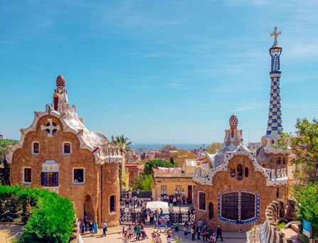 Der Park Güell mit seinen künstlerischen Gebäuden erstreckt sich vor der Skyline von Barcelona. Im Hintergrund das tiefblaue Meer und ein hellblauer Himmel.