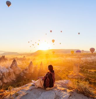 Eine Frau sitzt an einem Felsabsprung und blickt auf die Schlucht von Kappadokien. DIe Sonne geht gerade unter und taucht das gesamte Bild in ein orangenes Licht. Unzählige Heißluftballons schweben in der Luft.