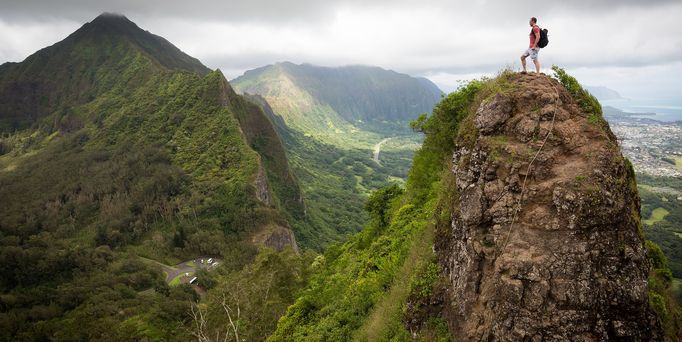 Ein Mann mit Rucksack steht einsam auf einem steil hervorstechendem Berg. Die Berglandschaft um ihn herum ist grün.