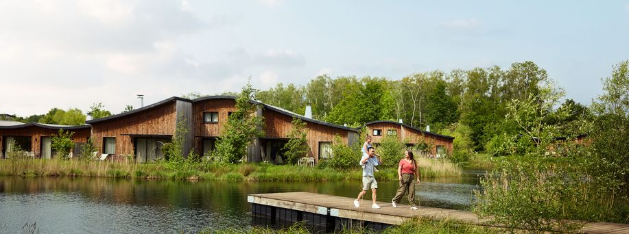 Eine dreiköpfige Familie läuft an einem See auf einem Steg in Richtung grüne Landschaft. Im Hintergrund direkt am See stehen moderne, naturbelassene Holzhäuser.