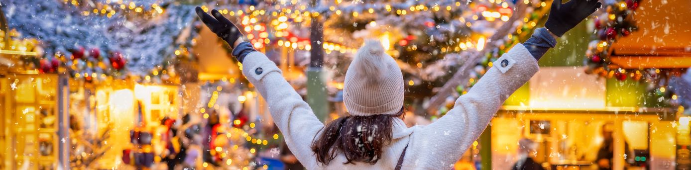 Eine Frau steht dem Betrachtenden mit dem Rücken zugewendet und armen in der Luft auf einem Weihnachtsmarkt. Sie trägt Mütze und Handschuhe und im Hintergrund sieht man viele gold-glitzernde Lichter.