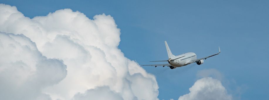 Ein weißes Flugzeug fliegt durch den blauen Himmel, welcher sich kontrastreich von den weißen puffrigen Wolken abhebt.