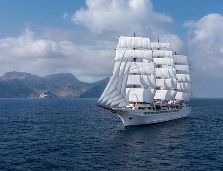 Ein riesiges weißes Segelschiff auf dem tiefblauen Meer. Im Hintergrund Berge.
