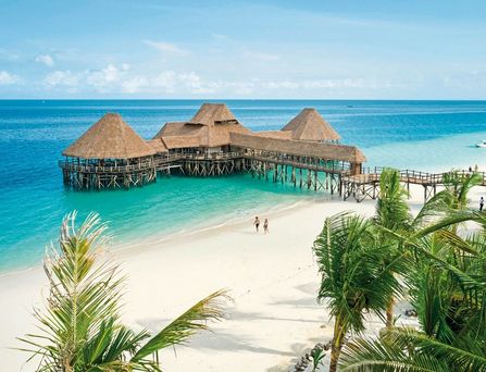 An einem menschenleeren Strand ragt ein Holzsteg ins türkis, blaue Wasser mit traditionellen Bungalows.