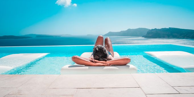Luxusurlaub: Eine Frau liegt auf einer Liege im Infinitypool mit Blick auf das Meer und die Berge