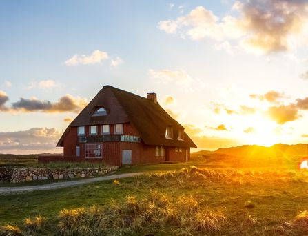 Ein großes rotes Haus mit Strohdach steht einsam in einer weiten, grünen Landschaft. Der Himmel ist von der untergehenden Sonne rosa-gold gefärbt und ganz rechts steht ein rot-weißer Leuchtturm.