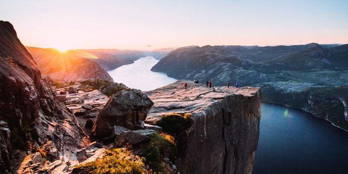 Die Sonne steht tief und taucht eine Berglandschaft, angrenzend an einen Fjord in orangenes Abendlicht. Einige Personen stehen an einem Abhang und genießen die Aussicht.