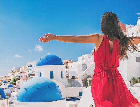 Eine brünette Frau in rotem Sommerkleid steht mit weit auseinandergebreiteten Armen vor Santorin. Ein Meer aus weißen Häusern mit blauen Dächern erstreckt sich vor dem blauen Meer.