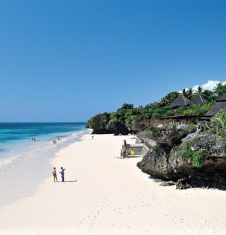 An einem weißen Sandstrand mit türkis-blauem Wasser laufen wenige Menschen entlang. Direkt am Strand grenzt eine Dschungellandschaft mit Bungalows.