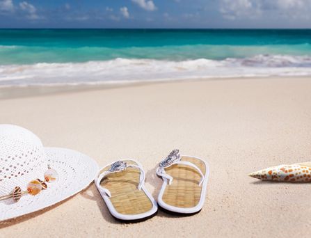 An einem Sandstrand liegen 2 weiße FlipFlops, ein weißer Sonnehut und eine Muschel.