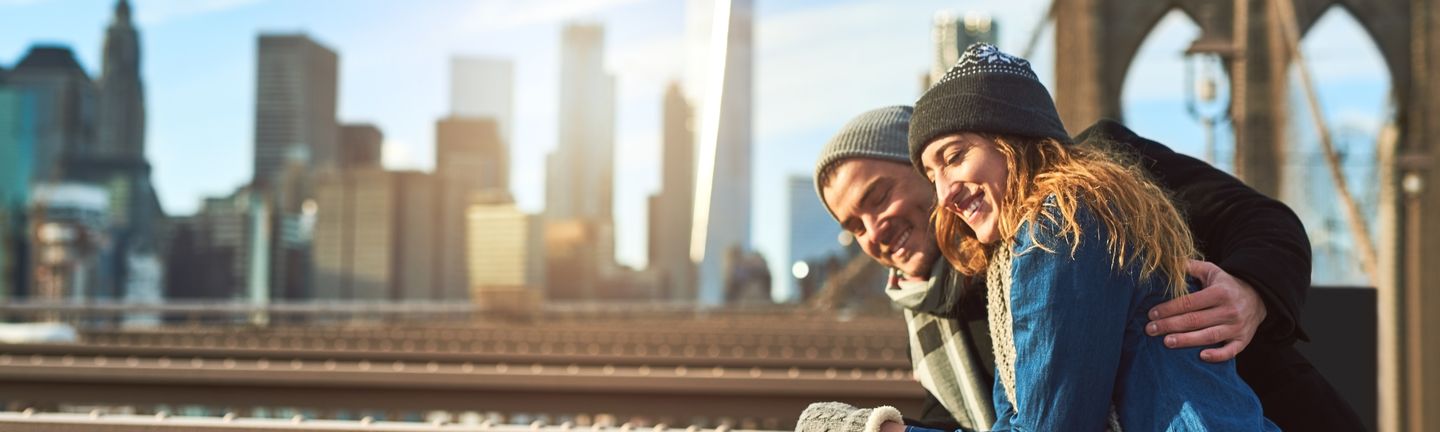 Ein junges Paar steht lachend in Winterklamotten auf der New Yorker Brooklynbridge.