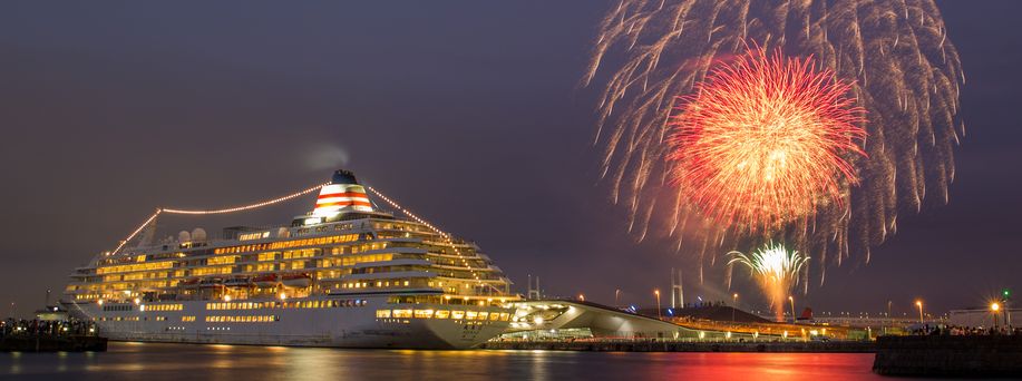 Ein beleuchtetes Kreuzfahrtschiff befindet sich an einem Hafen. Es ist dunkel und im Hintergrund leichtet das Feuerwerk.