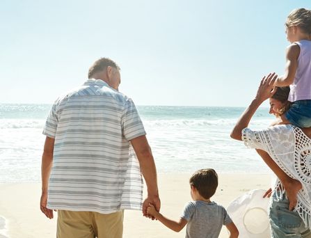 Eine Familie, bestehend aus einer Frau, 2 Kindern im Alter von ca. 4 bis 6 Jahren und einem älteren Mann, laufen an einem Sandstrand aufs Meer zu. Das Mädchen sitzt bei der Frau auf den Schultern und der Junge geht an der Hand des Mannes.