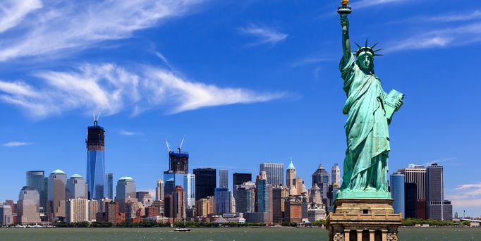 Die Freiheitsstatue vor dem Hudson River und der Skyline von News York mit vielen Wolkenkratzern unter blauem Himmel.