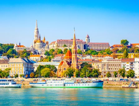 In strahlenden Farben erstreckt sich die Skyline von Budapest mit Gebäuden und Kirchen am Ufer eines Flusses, auf welchem 2 Schiffe fahren.