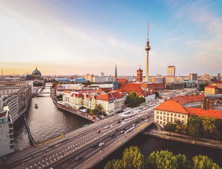 Eine befahrene Brücke führt in die Berliner Stadtmitte mit rotem Rathaus, dem Fernsehturm und dem Berliner Dom. Der Himmel ist blau mit gelb-weißem Licht durchzogen.
