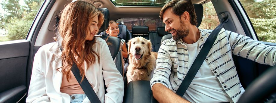 Eine junge dreiköpfige Familie mit Golden Retriever sitzt im Auto. Alle schauen den Hund an und lächeln.