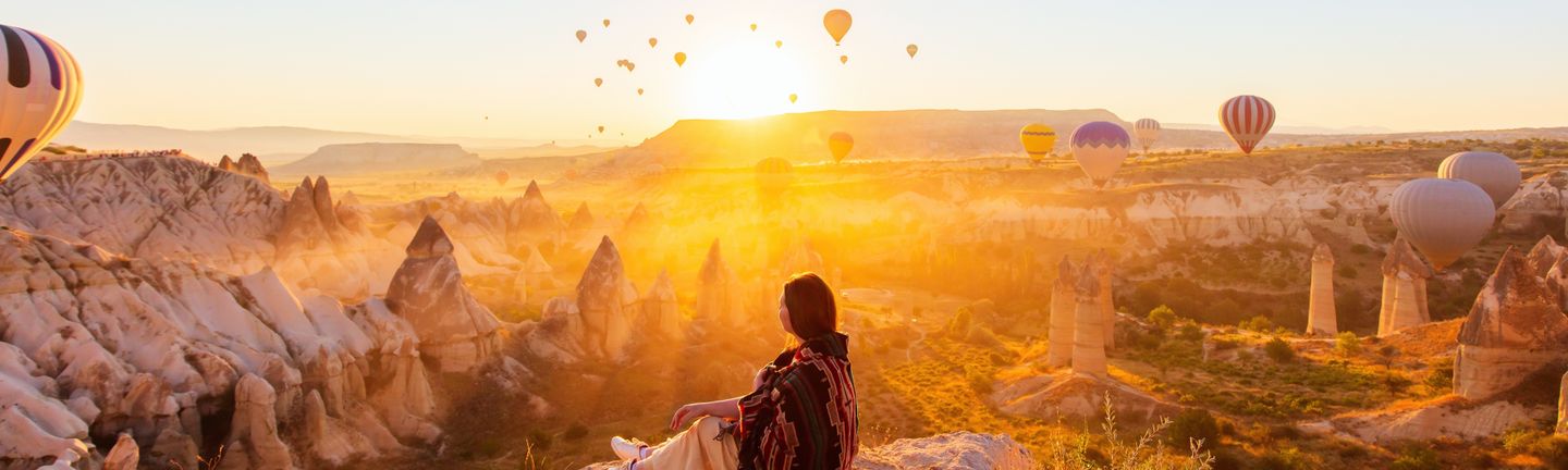 Eine Frau sitzt an einem Felsabsprung und blickt auf die Schlucht von Kappadokien. DIe Sonne geht gerade unter und taucht das gesamte Bild in ein orangenes Licht. Unzählige Heißluftballons schweben in der Luft.