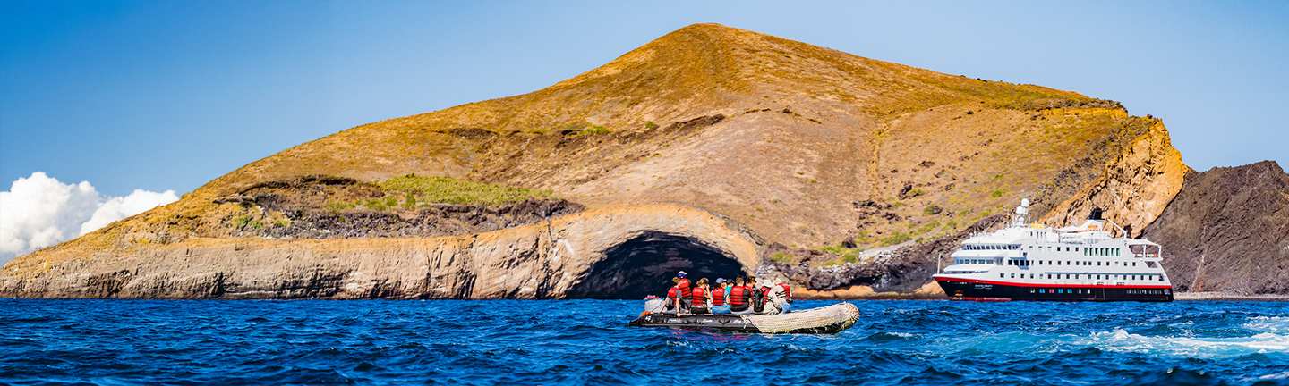 Eine Frau steht im Sommerkleid mit Sonnenhut lachend in einem Weinanbaugebiet. Sie hält ein Weinglas vor sich in welches gerade Rotwein geschüttet wird. Im Hintergrund steht ein Mann, welcher herzhaft lacht, ebenfalls mit einem Rotweinglas in der Hand.