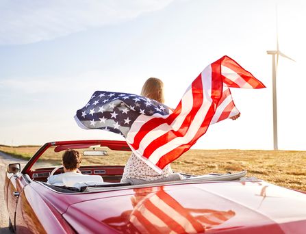 Ein Pärchen fährt in einem Oldtimer Cabrio eine Landstraße entlang und die Beifahrerin schwingt eine USA Flagge im Wind hinter sich mit.