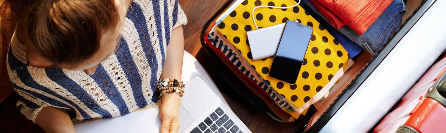Eine Frau sitzt mit Laptop und Klemmbrett mit Stift und Papier auf dem Boden neben ihrem aufgeklappten Koffer. In diesem befinden sich neben Klamotten auch ein Handy und eine Powerbank.