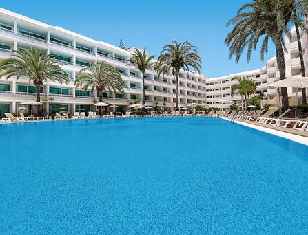 Direkt am Meer liegt eine Hotelanlage mit einer Poollandschaft zwischendrin immer wieder grüne Bäume und viele Sonnenschirme.