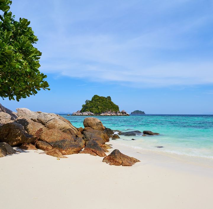 Ein feinsandiger, weißer Strand mit großen Steinen, welche ins türkisblaue, klare Meer ragen. Im Hintergrund eine kleine Insel.